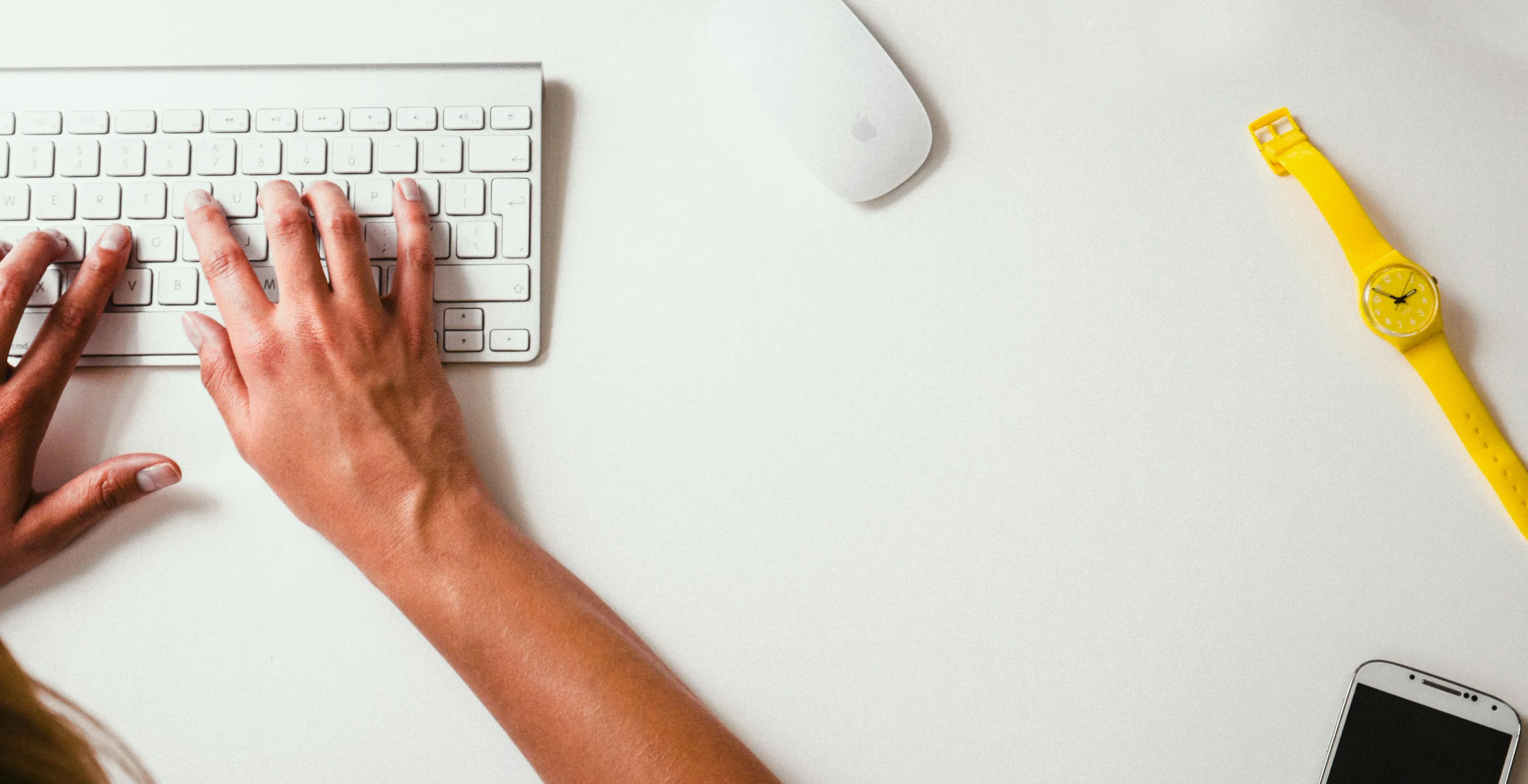 Hands typing on a white keyboard, with a yellow watch and a phone on the desk