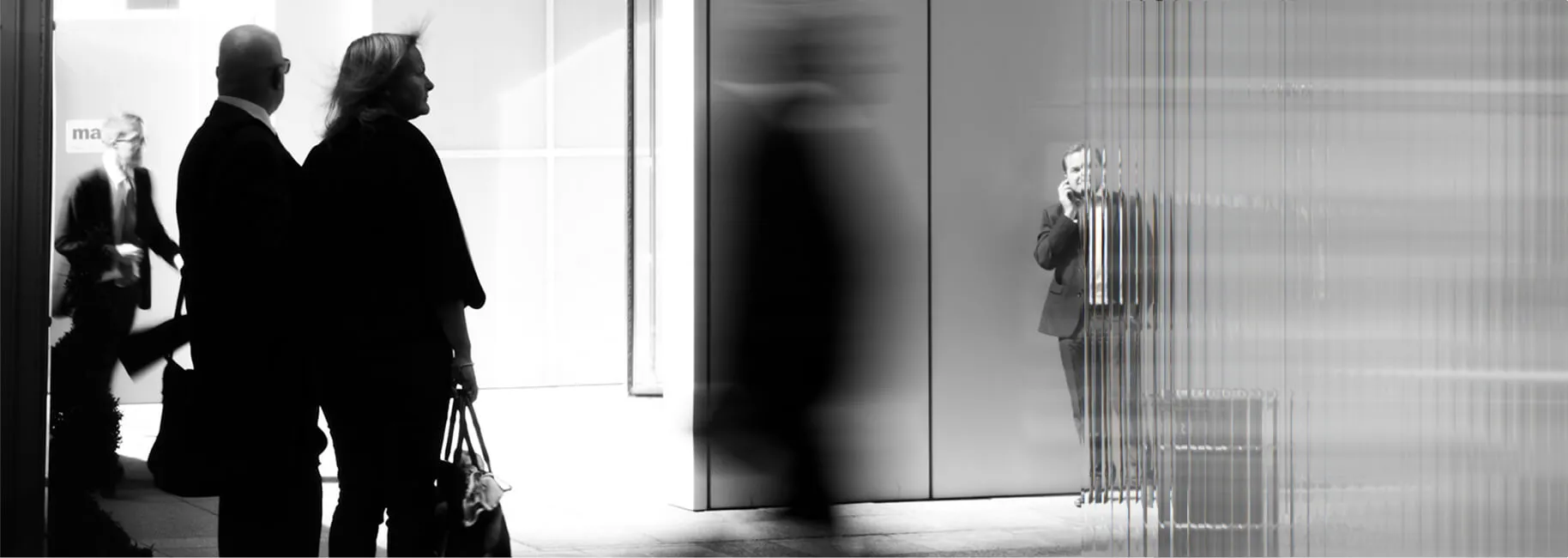 Black and white street scene of people outside a glass office building, with a man on the phone behind the facade